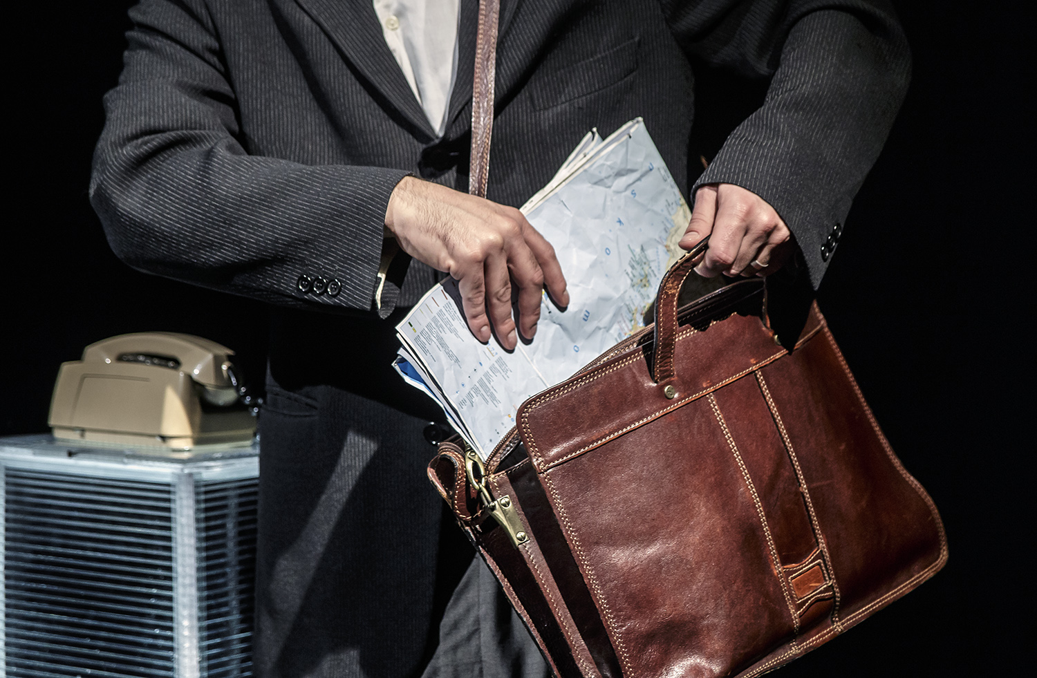 Dettaglio di uomo in giacca e pantaloni, ripone una cartina dell'Istria in una borsa di cuoio appesa alla spalla. Dietro un vecchio telefono a disco.