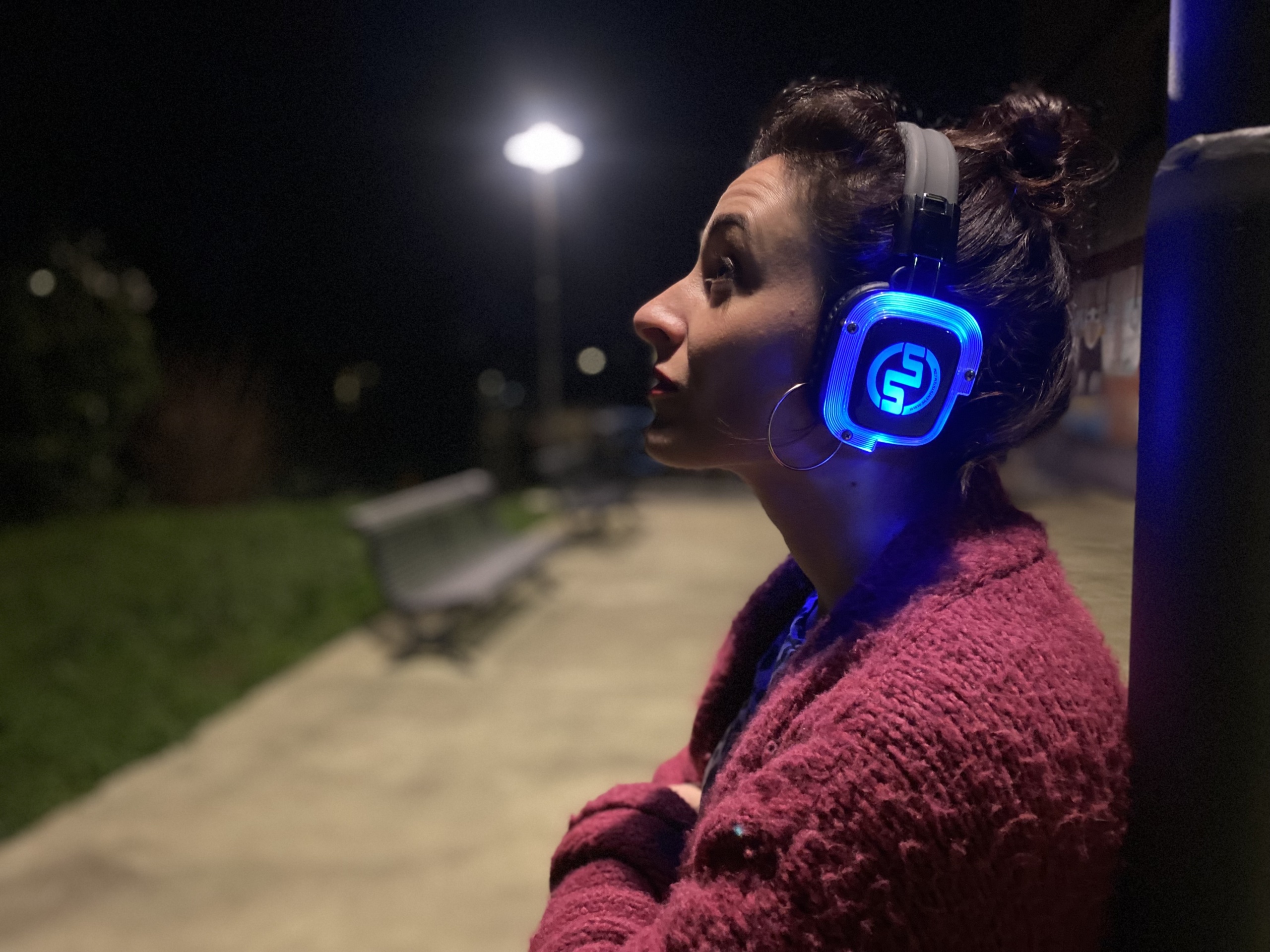 Sullo sfondo di un parco urbano illuminato dalla luce di un lampione, una donna scruta la città di notte. Ha le braccia incrociate e indossa cuffie audio.