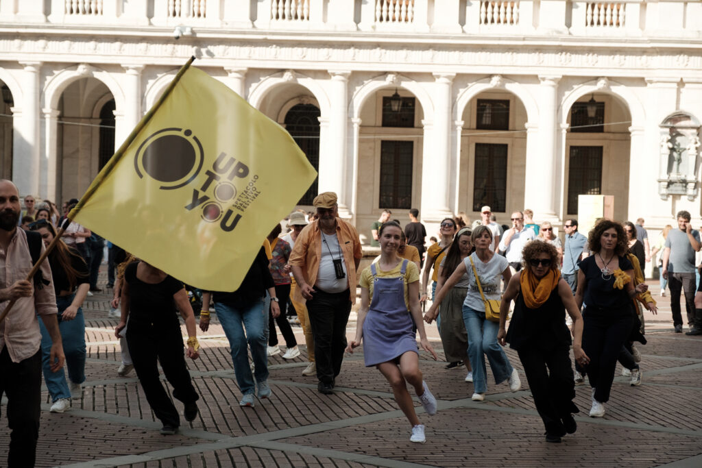 Un gruppo di persone danza in una piazza cittadina. Sventola una bandiera con scritto Up To You spettacolo dal vivo festival